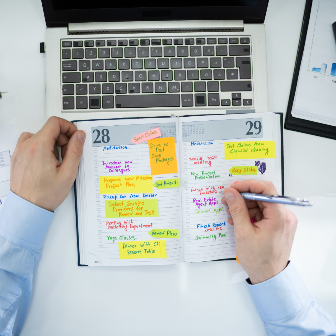 agenda with many tasks in different colors lying next to the laptop, hands are holding the agenda and a pen symbolizing prioritization of tasks