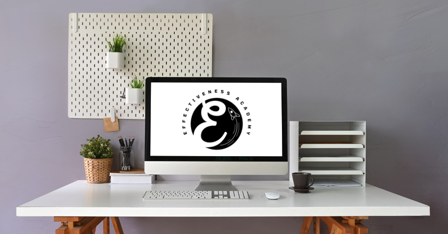 A minimalist yet inviting workspace featuring a modern stationary PC with the Effectiveness Academy logo on the monitor, signifying the 'Planify' digital tool. The PC is placed on a pristine white desk alongside a neat paper holder, promoting organization and efficiency. A wall-mounted board with two small potted plants adds a touch of greenery, offering a serene working atmosphere. This picture subtly encourages the viewer to utilize 'Planify' for a streamlined, effective approach to planning tasks.